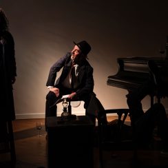 Na scenie po lewej stronie stoi kobieta, obok mężczyzna siedzi na stołku, uśmiecha się do kobiety, z prawej strony stoi fortepian, na nim butelka z winem i kieliszki.